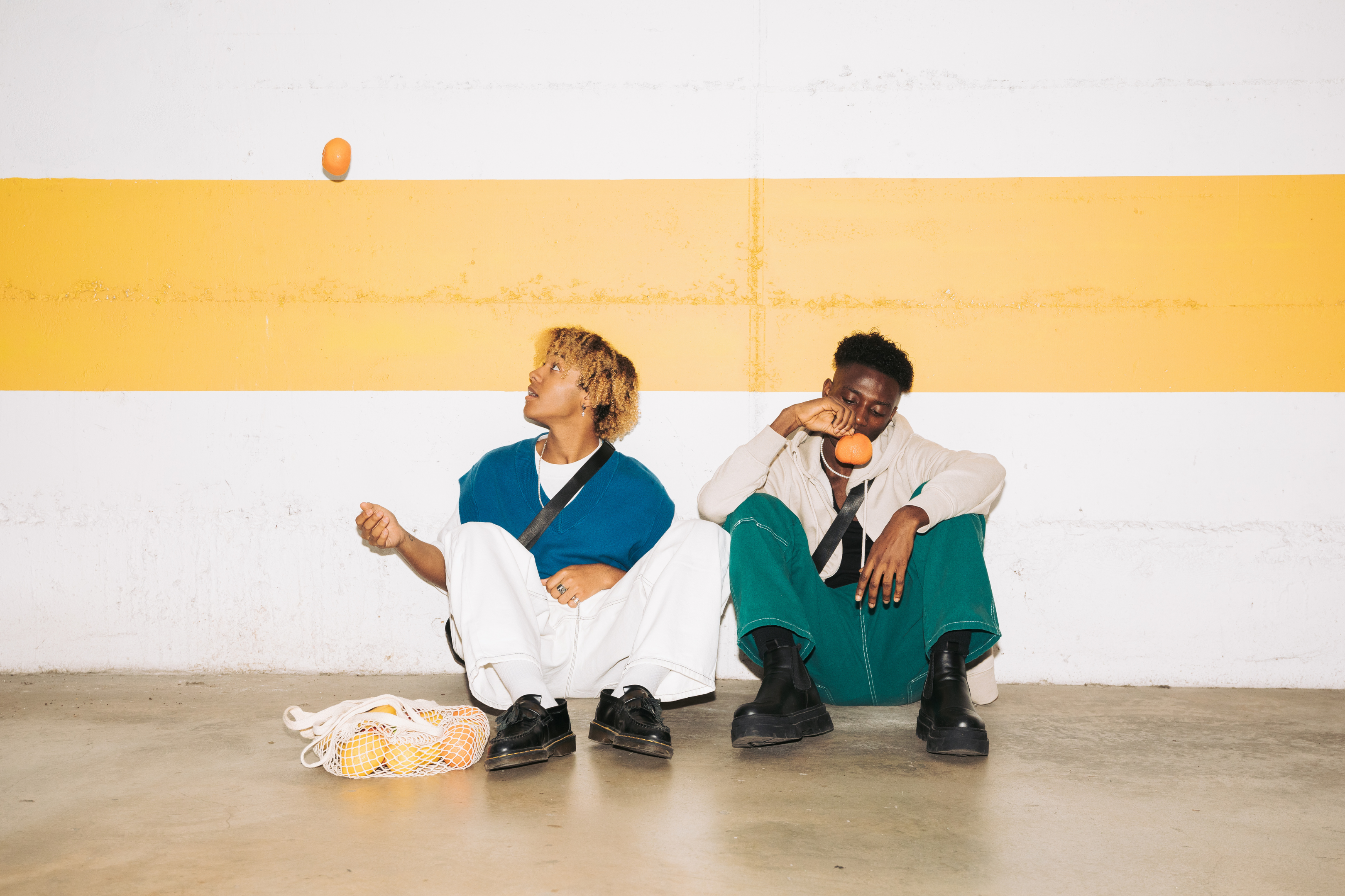 Two people sitting on the floor with oranges against a yellow and white wall.